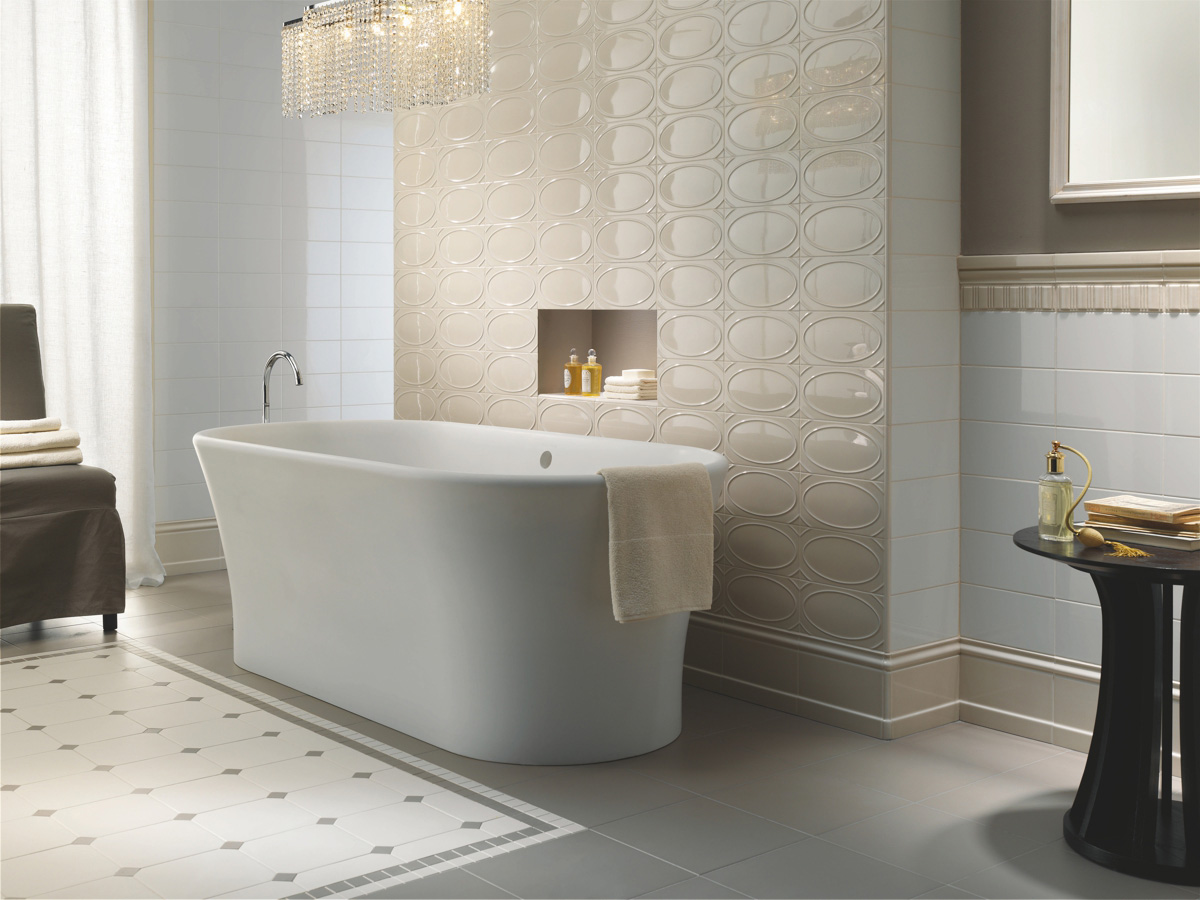 Luxusní koupelna s obklady Ceramiche Grazia Formae s lesklými béžovými oválnými reliéfy na zdi za vanou. Ostatní stěny jsou obloženy bílými a béžovými obklady. Na podlaze je šedá dlažba s bílým a tmavým bordurou. Uprostřed stojí bílá volně stojící vana. Prostor doplňuje křišťálový lustr a elegantní nábytek.