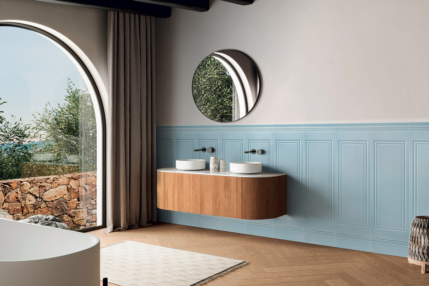 Moderní koupelna s modrými obklady Elegance 2.0 s reliéfním vzorem, které pokrývají dolní část stěny. Nad nimi je světlá stěna. Místnost má dřevěnou parketovou podlahu, závěsnou dřevěnou skříňku se dvěma bílými umyvadly a kulaté zrcadlo. Dominantou je velké obloukové okno s výhledem do krajiny.